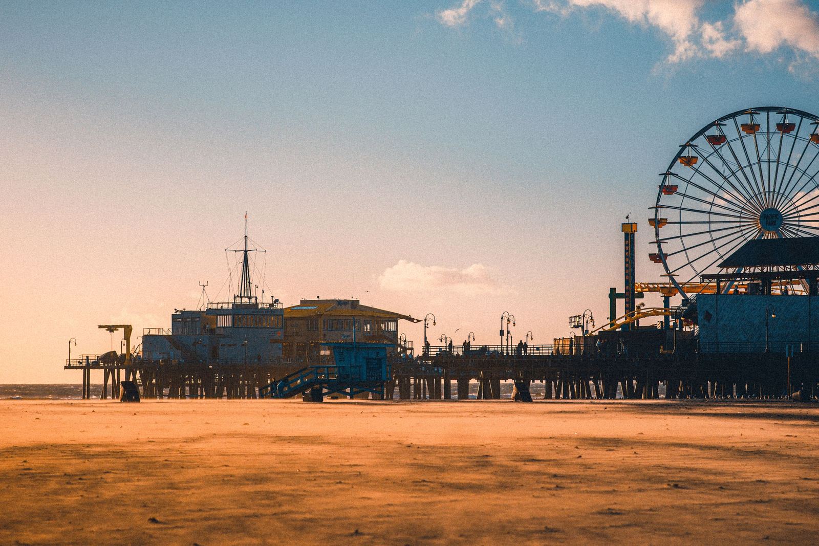 Santa Monica Pier at dusk, wind blowing sand across an empty beach, the ferris wheel silhouetted under a warm sky