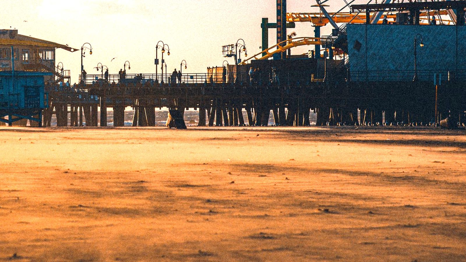 Santa Monica Pier at golden hour seen from the beach, the deck, lamps, people and rides all resting on the pilings below