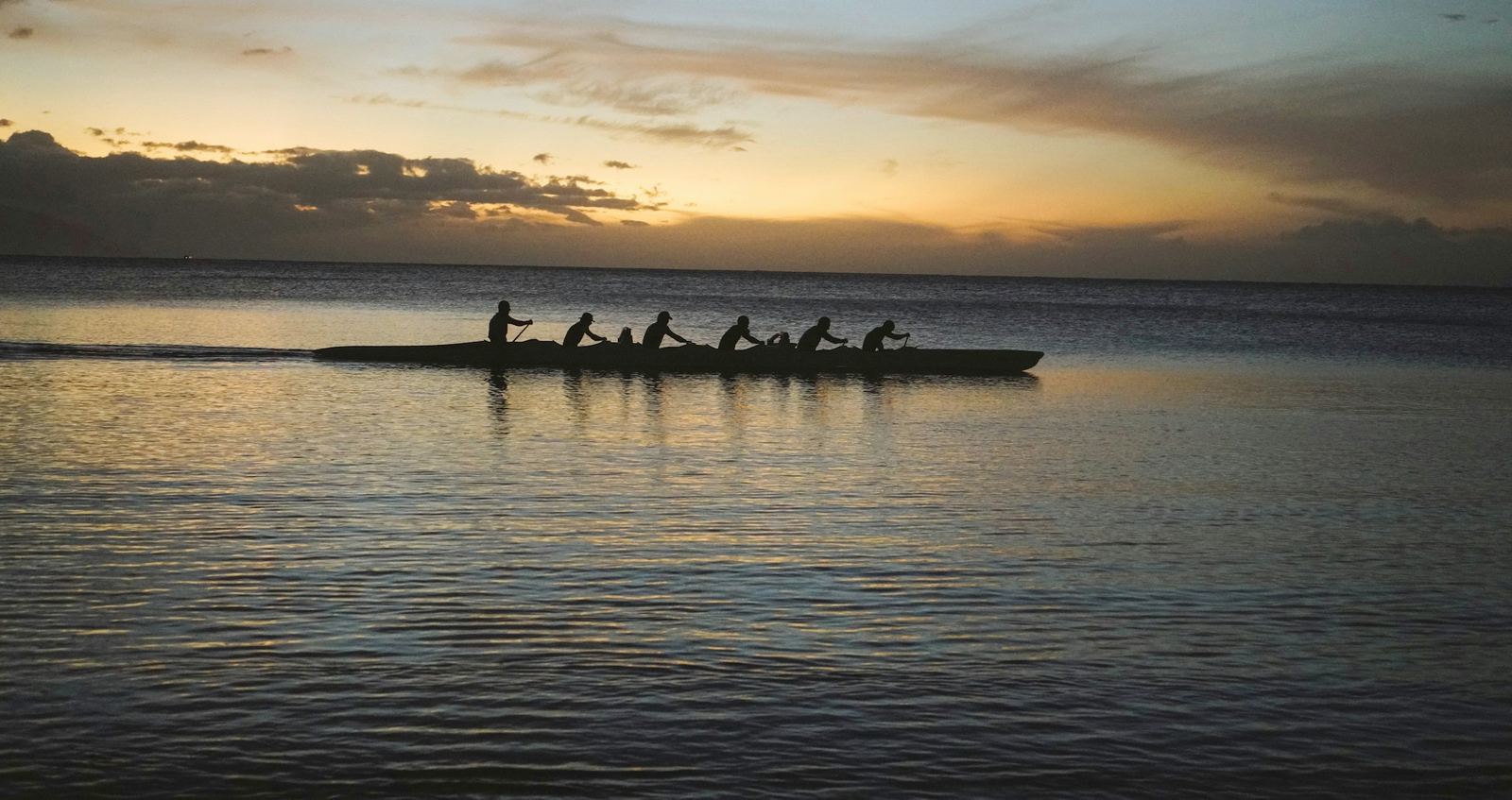 An outrigger canoe crew in silhouette, paddling in unison across calm water at sunset