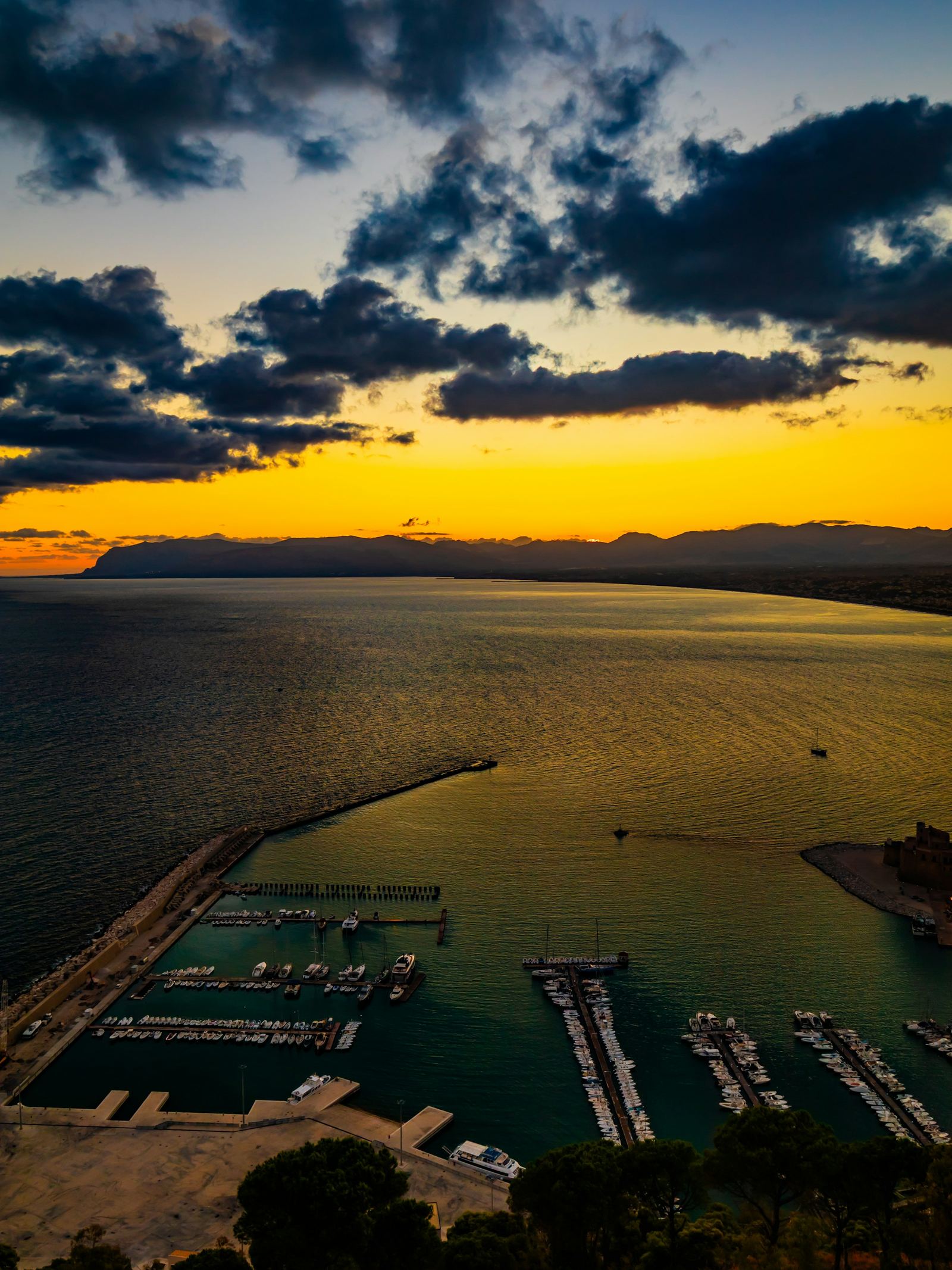 Elevated view over a harbor and its marina at golden hour, under an amber sky and dark clouds
