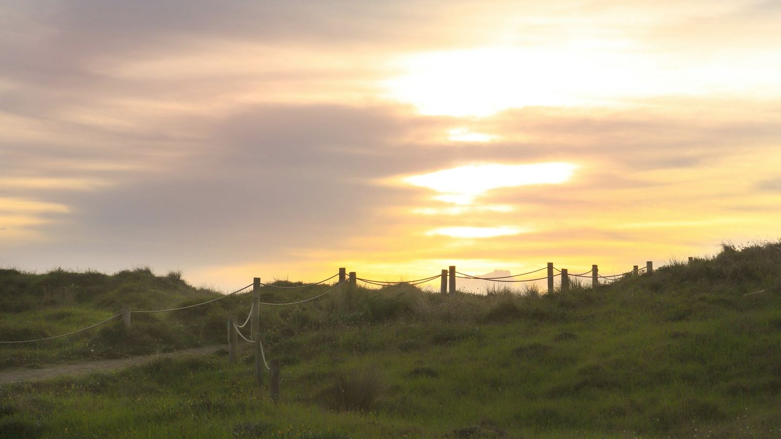 A rope-and-post fence running along a grassy coastal dune beneath a golden sunset sky