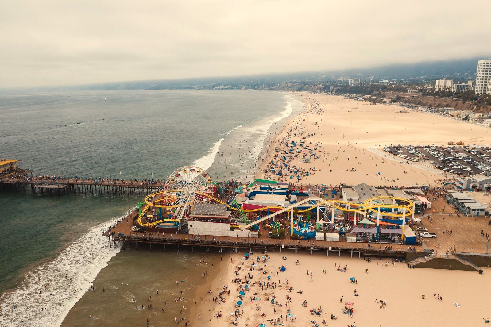 Aerial view of the Santa Monica Pier, the ferris wheel and roller coaster running on the boardwalk below, the coastline curving north behind